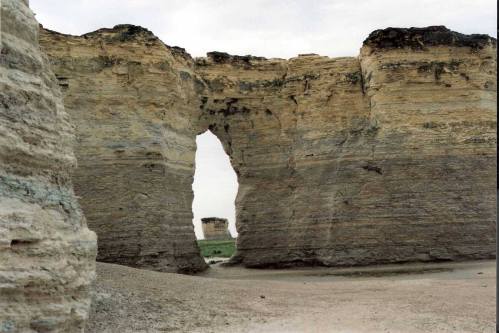 A natural rock formation with a large archway, surrounded by sandy terrain and a cloudy sky.