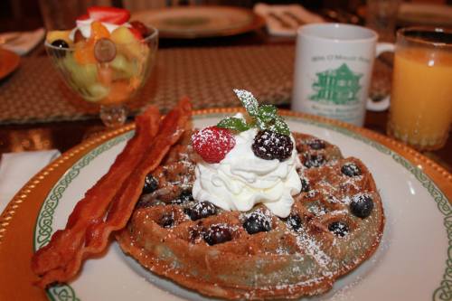 A plate with a waffle topped with whipped cream, berries, and powdered sugar, alongside bacon and a fruit cup.