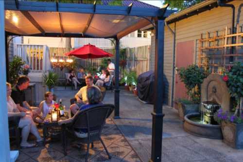 A cozy outdoor patio with people socializing, surrounded by plants, a fountain, and warm lighting under a canopy.