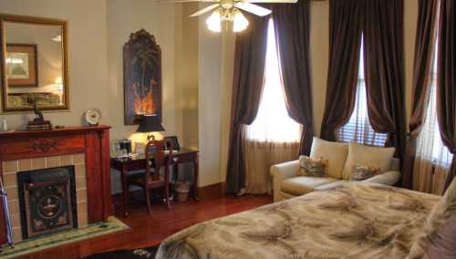 Cozy bedroom with a fireplace, desk, and large windows, featuring elegant curtains and warm wooden accents.