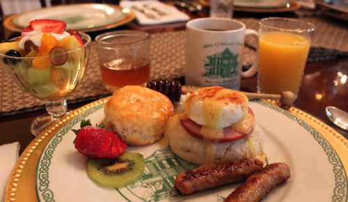 A plate with eggs Benedict, sausage, a biscuit, and fresh fruit, accompanied by drinks and a decorative cup.