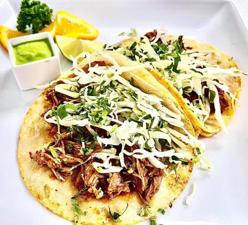 Two tacos filled with shredded meat, topped with cabbage and herbs, served with lime, orange, and a small dish of green sauce.