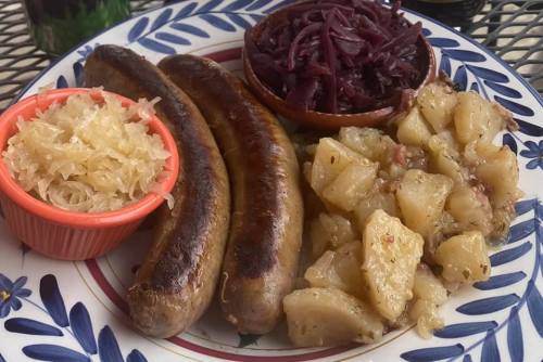 A plate featuring three grilled sausages, sauerkraut, red cabbage, and seasoned potatoes, all beautifully arranged.