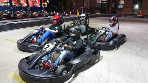 Five people in go-karts, wearing helmets, ready to race on an indoor track.