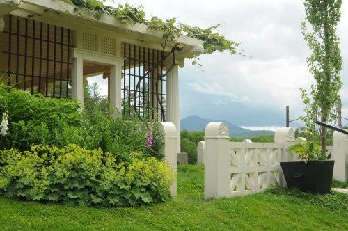 A charming garden with lush greenery and a white gate, set against a backdrop of mountains and cloudy skies.
