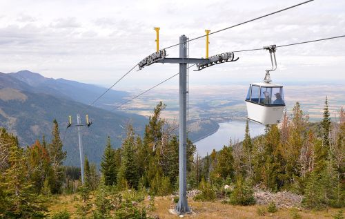 A gondola lift ascends a mountain, overlooking a valley and river, surrounded by trees and distant mountains.
