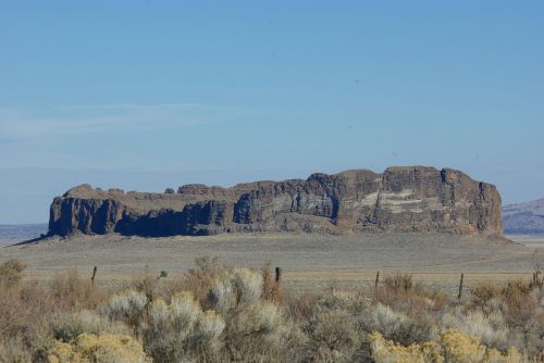 A flat-topped rock formation rises above a desert landscape under a clear blue sky. Sagebrush is visible in the foreground.