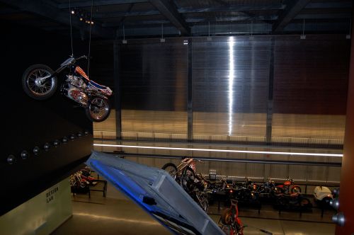 A motorcycle hangs from the ceiling in a modern exhibit space, with various bikes displayed below.