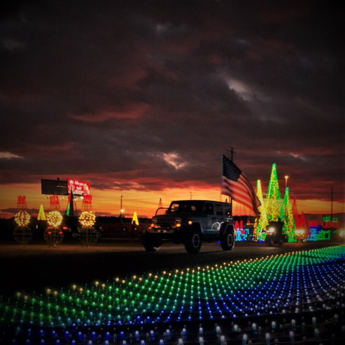 A colorful display of holiday lights with a sunset backdrop and a vehicle driving through the scene.