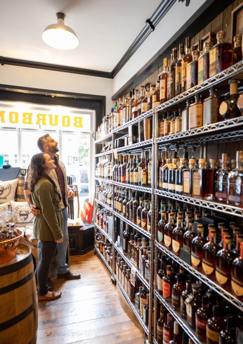 A couple admires a wall of whiskey bottles in a cozy shop, with wooden barrels and a bright window in the background.