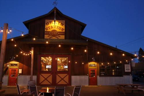 A rustic brewery building at dusk, adorned with string lights and a fire pit in front, creating a cozy atmosphere.