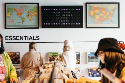 Two people browse records in a store, with maps and a sign about Rough Trade displayed on the wall above.