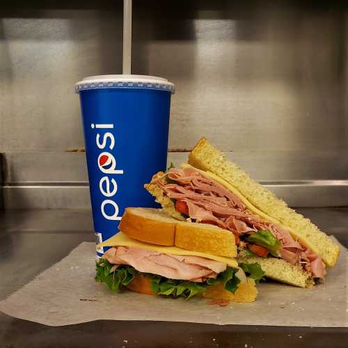 A sandwich with layers of turkey, ham, and lettuce beside a blue Pepsi cup on a countertop.