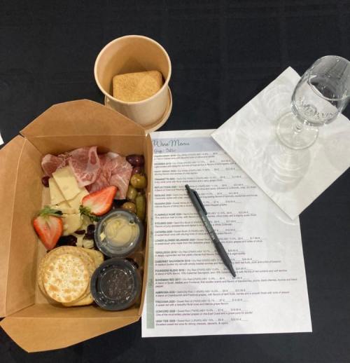 A takeout box with cheese, meats, fruit, and crackers, alongside a menu, glass, and pen on a black table.