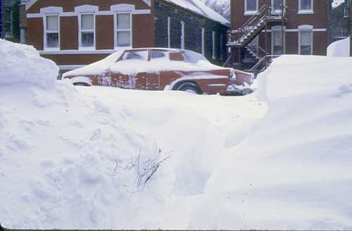 A red car partially buried in deep snow, with snowdrifts on either side and brick buildings in the background.