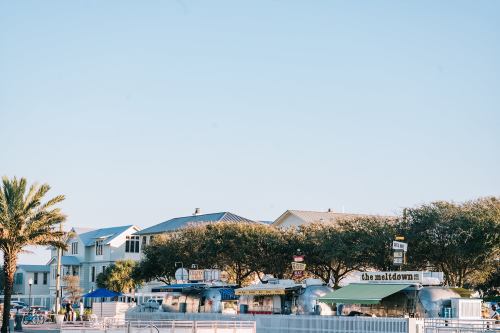 A beachside scene featuring food stalls, palm trees, and nearby houses under a clear blue sky.