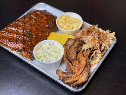 A metal tray with barbecue ribs, pulled pork, coleslaw, cornbread, and crispy bacon, served with two side cups.