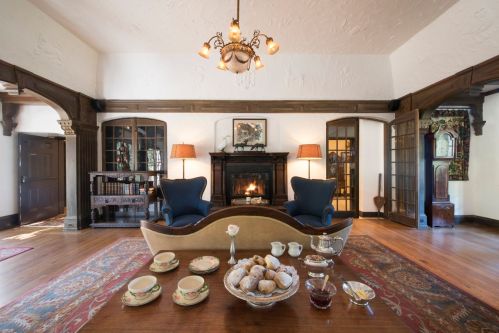 Cozy living room with a fireplace, elegant furniture, and a table set with tea and pastries. Warm, inviting atmosphere.