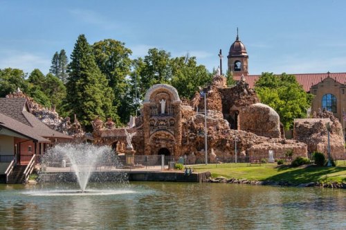 A scenic view of a historic stone structure by a lake, with a fountain and trees in the background under a clear blue sky.
