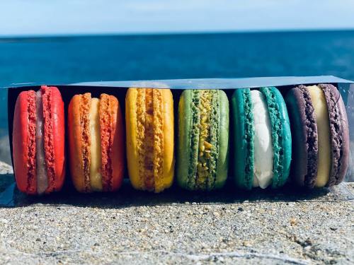 A colorful assortment of macarons arranged in a row on a sandy surface, with a blue ocean in the background.
