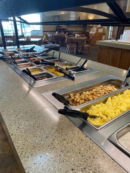 A buffet table with various food trays, including scrambled eggs, potatoes, and meats, in a dining area.