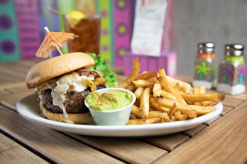 A juicy burger with cheese and an umbrella, served with fries and guacamole on a wooden table.