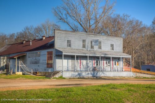 A weathered two-story building with a porch, surrounded by trees and a gravel path.