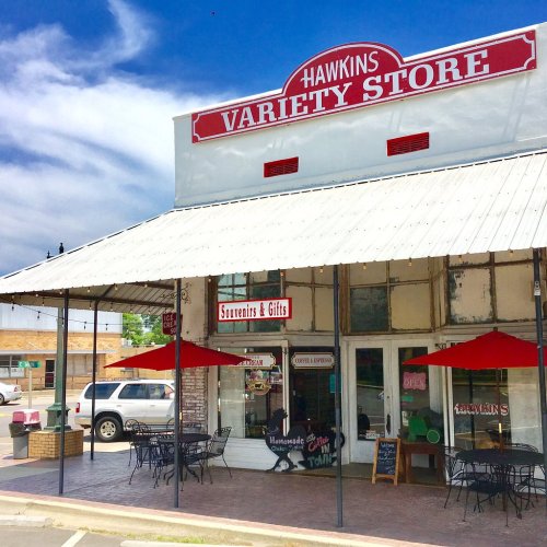 A colorful variety store with outdoor seating, featuring a sign that reads "Hawkins Variety Store" under a blue sky.