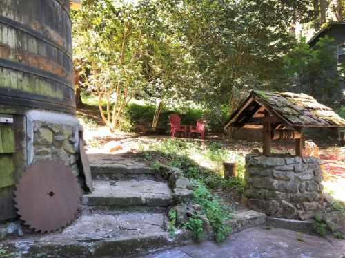 A rustic scene featuring a wooden barrel, a stone well, and two red chairs in a lush, green garden.