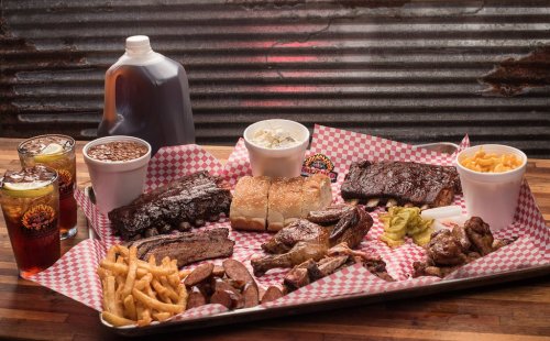 A platter of assorted barbecue meats, sides, and drinks, featuring ribs, sausage, fries, and coleslaw on a checkered tablecloth.