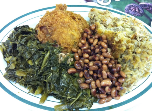 A plate of Southern-style food featuring fried chicken, collard greens, black-eyed peas, and cornbread stuffing.