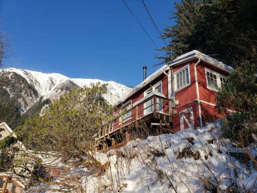 A red house on a snowy hillside, surrounded by greenery, with mountains in the background under a clear blue sky.