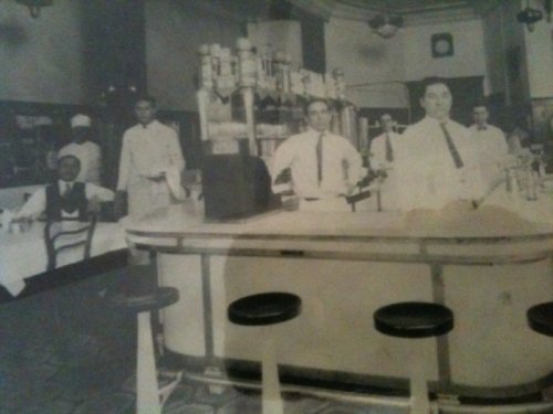 Black and white photo of a vintage diner with staff behind the counter and customers seated at tables.