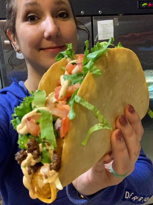 A person holds a large taco filled with beef, lettuce, tomatoes, and sauce, smiling at the camera.