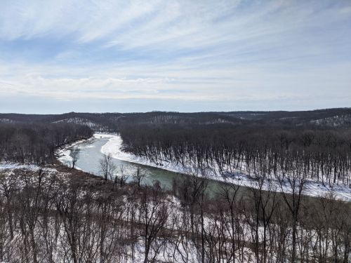 A winding river flows through a snowy landscape, surrounded by bare trees and rolling hills under a cloudy sky.