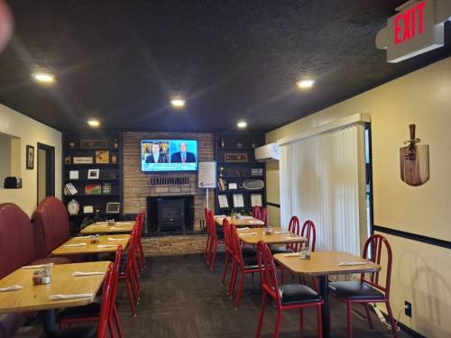 Cozy restaurant interior with red chairs, wooden tables, a TV on the wall, and a fireplace. Exit sign visible.