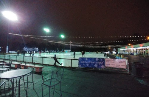 A nighttime ice skating rink with skaters, string lights, and a festive atmosphere. Tables are set up nearby.