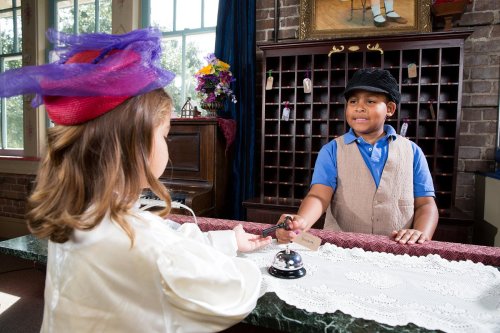A young girl in a hat interacts with a boy at a reception desk, with a bell and decorative items in the background.