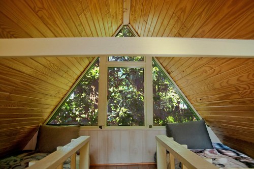 Cozy attic space with wooden walls, two beds, and a large triangular window overlooking greenery.