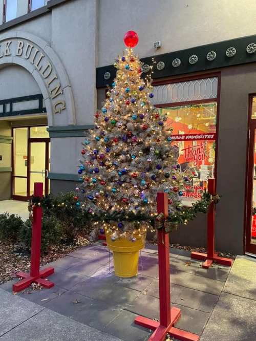 A decorated Christmas tree with colorful ornaments and lights, placed outside a building, surrounded by festive garland.