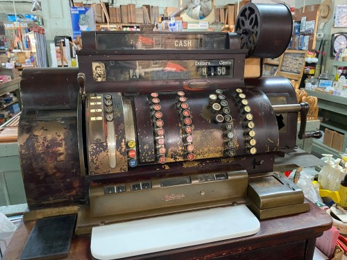An antique cash register with a wooden base, buttons, and a display showing $48.50 in a vintage shop setting.