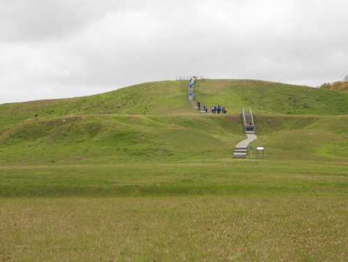 A grassy hill with a winding path leading up, where people are walking towards the top under a cloudy sky.