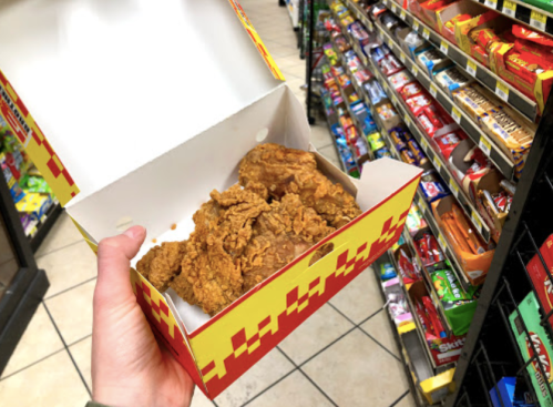 A hand holds a box of fried chicken in a convenience store aisle filled with snacks and candy.
