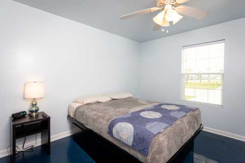 A simple bedroom with a bed, blue walls, a ceiling fan, a nightstand, and a window with blinds.