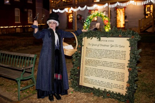 A woman in period costume holds a candle holder and a basket, standing beside a festive sign in a decorated outdoor setting.