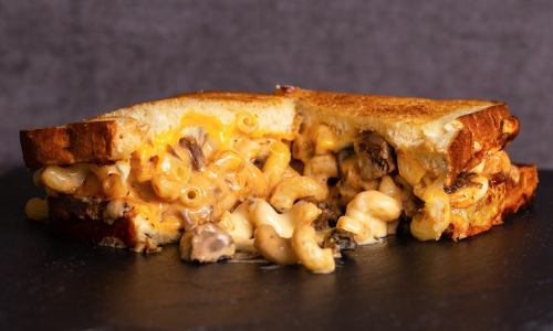 A grilled cheese sandwich filled with creamy macaroni and cheese, featuring melted cheese and mushrooms, on a dark background.