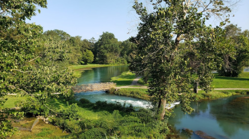 A serene landscape featuring a river, a small bridge, and lush greenery under a clear blue sky.