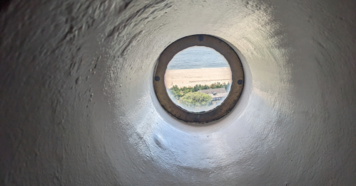 View through a circular opening, showing a beach and water beyond, framed by a textured white wall.