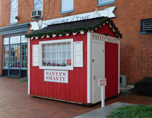 A festive red wooden booth labeled "Santa's Shanty," decorated with greenery, set against a brick building backdrop.