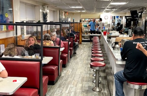 A diner interior with red booths, a long counter, and patrons enjoying meals, separated by clear partitions.